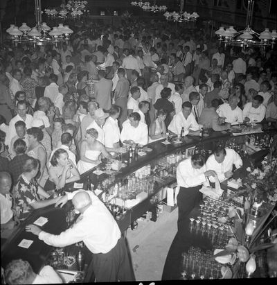 The Showboat's curved bar packed shoulder to shoulder on opening day in September 1954, white-shirted bartenders working under lamp chandeliers