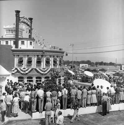 Black-and-white photograph of the Showboat's opening ceremony on September 3, 1954: a large crowd around the flag-bunted riverboat prow, a speaker at a lectern, parasols, 1950s cars, and empty desert with a Coca-Cola billboard beyond